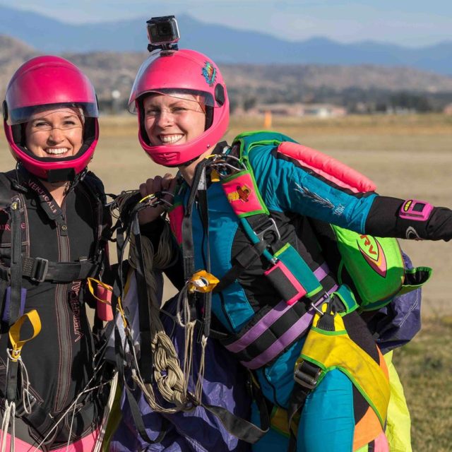 Two female skydivers with pink helmets and colorful rigs stand and pose for the camera