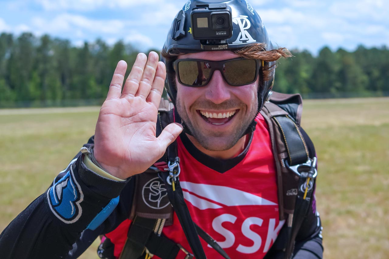 Smiling skydiver with a black helmet and red shirt mimes a high five