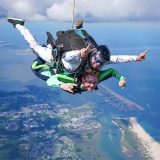 Tandem skydiving student and instructor smiling while in freefall over a beautiful coastline