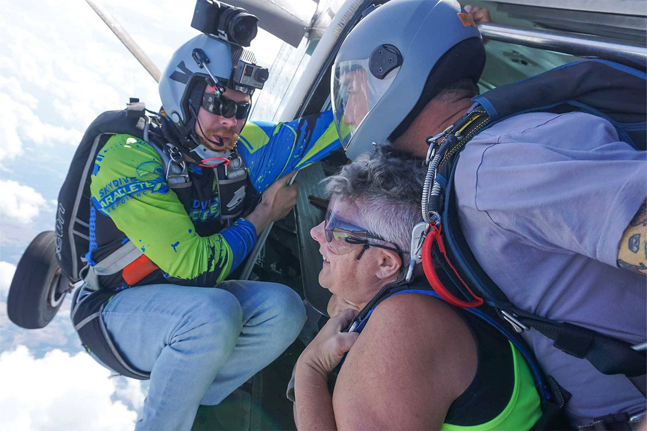 Videographer perched in the door of an airplane capturing a tandem skydiving student and instructor as the exit